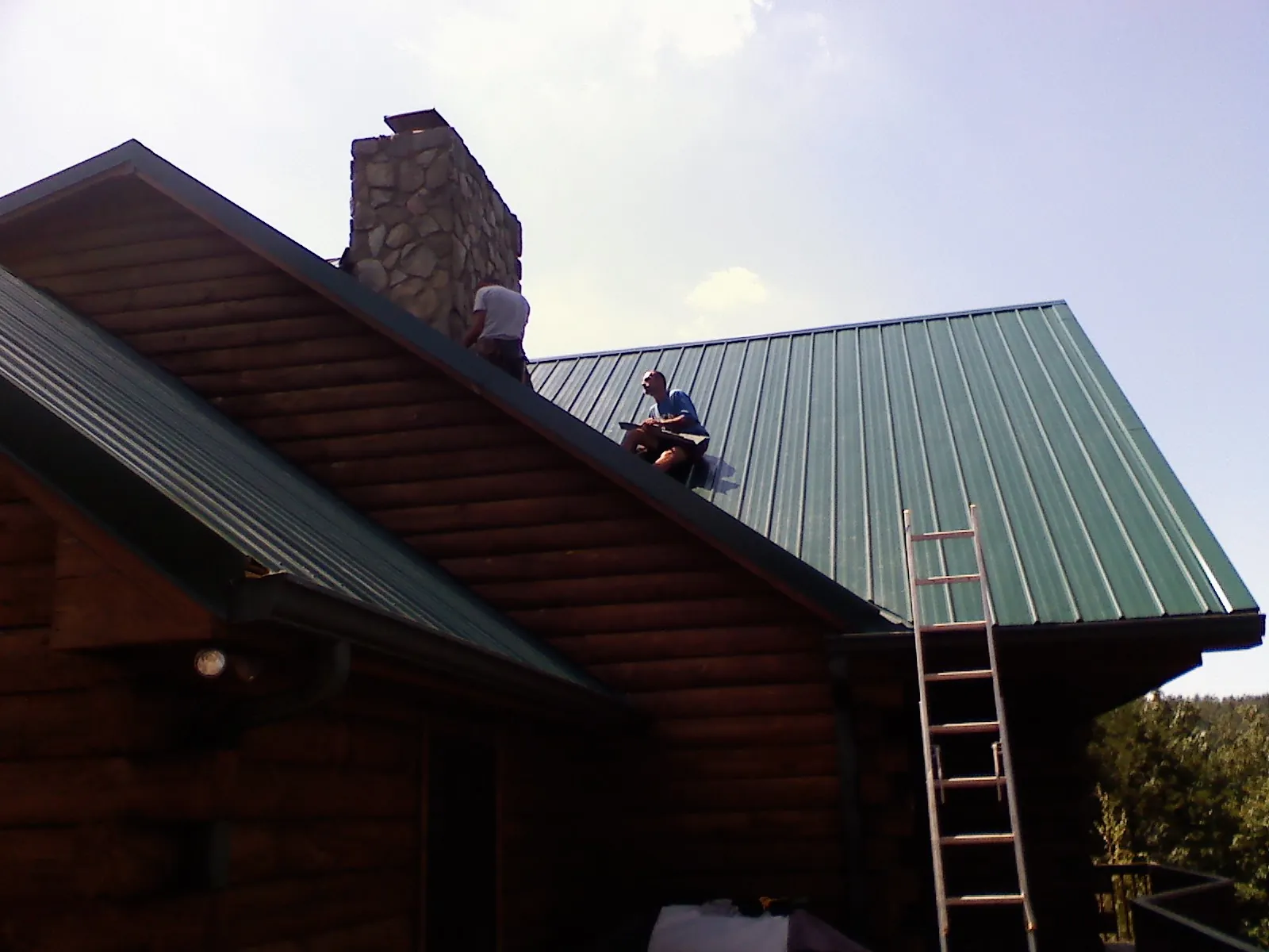 Roofers installing green metal panels beside a stone chimney on a log home in Egg Harbor